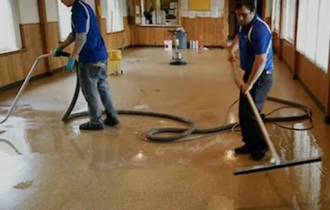 Cleaning and restoration services workers in blue uniforms using hoses and squeegees to remove water from a glossy hallway floor.